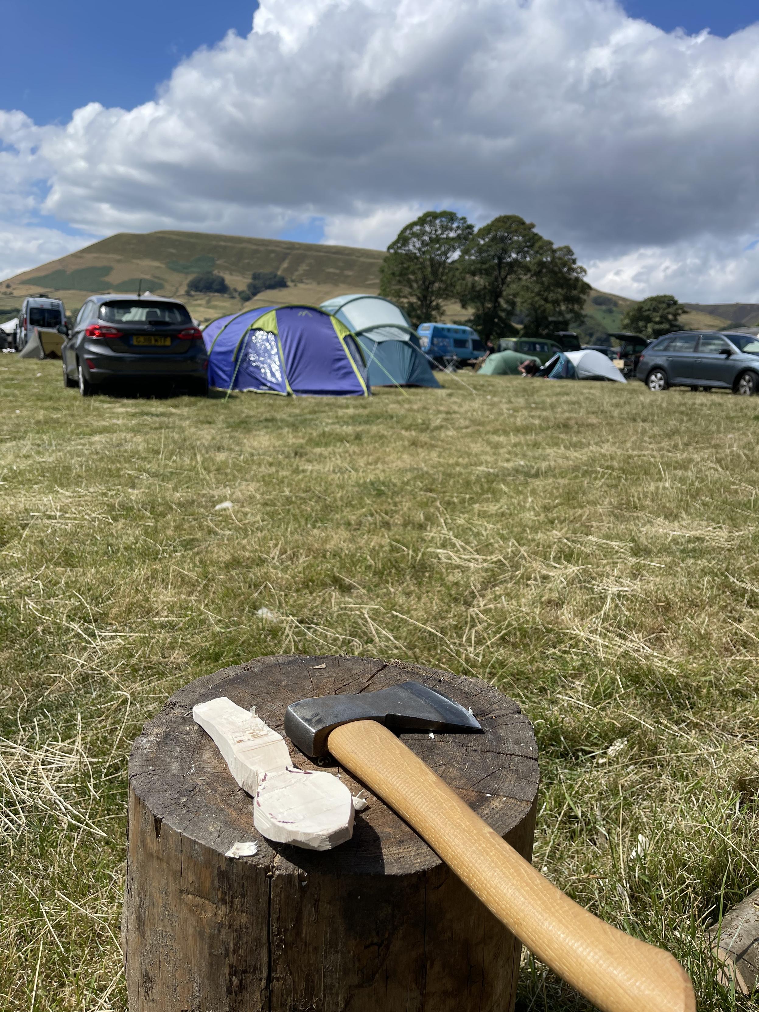 An axe on wooden block next to a rough carved spoon, with tents and the hills of the Peak District in the background.