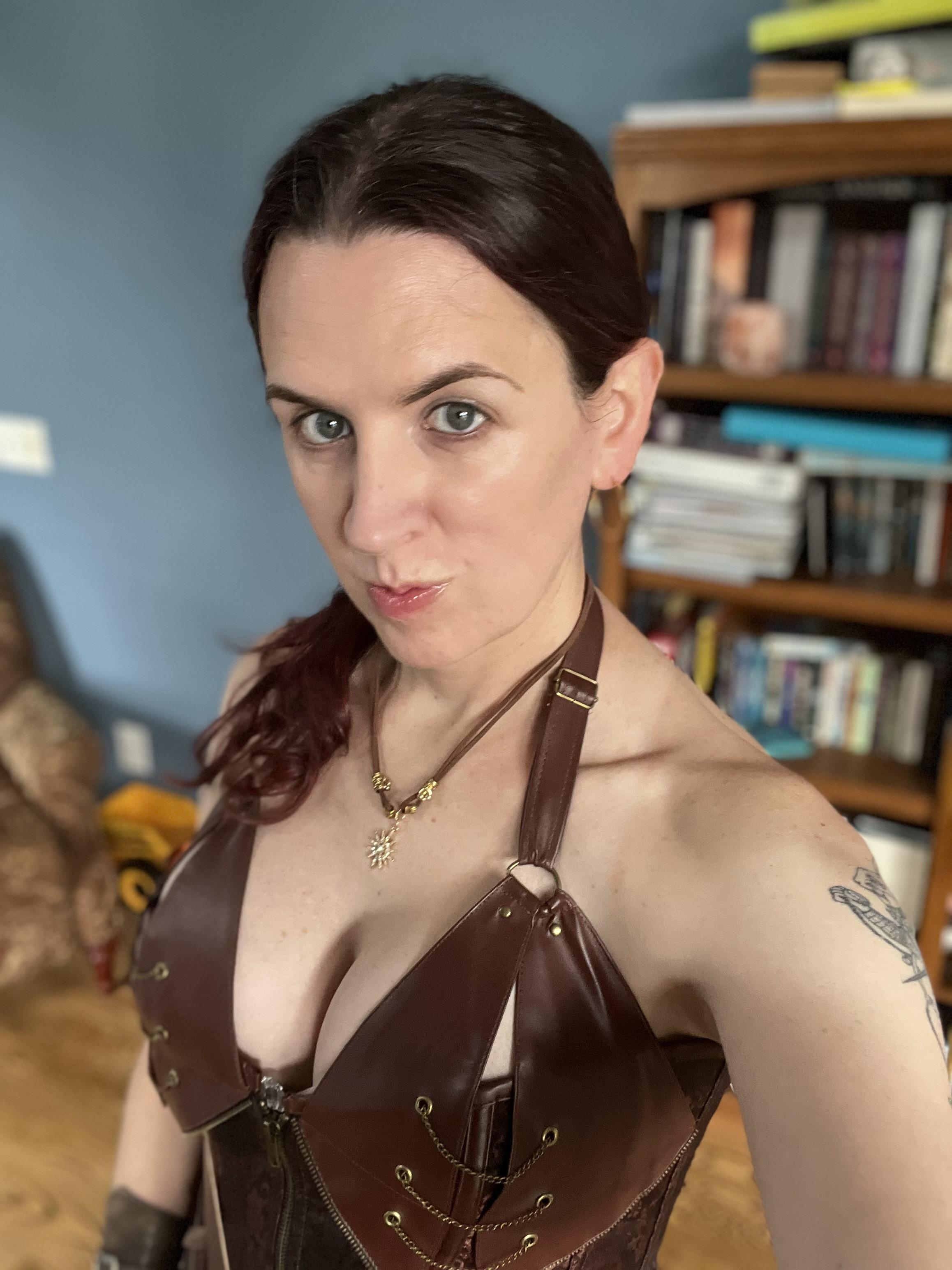 A high-angle selfie of a light-skinned woman with dark reddish-brown hair tied back. She wears a brown leather-look, halter-neck corset top with decorative gold chain accents and a matching sun-pendant necklace. She has a tattoo on her upper arm and poses toward the camera.