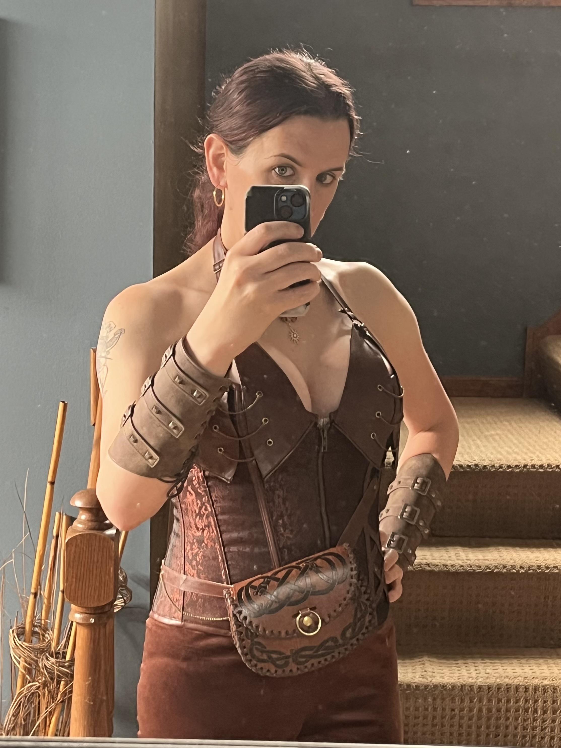 A front-facing mirror selfie of a light-skinned woman with dark hair tied back in a ponytail. She is wearing a dark brown and rust-colored leather-style fantasy corset top with lacing details, matching dark brown bracers on both forearms, and a brown leather pouch at her waist featuring dark Celtic knotwork details. She holds a black smartphone up in front of her face to take the photo.