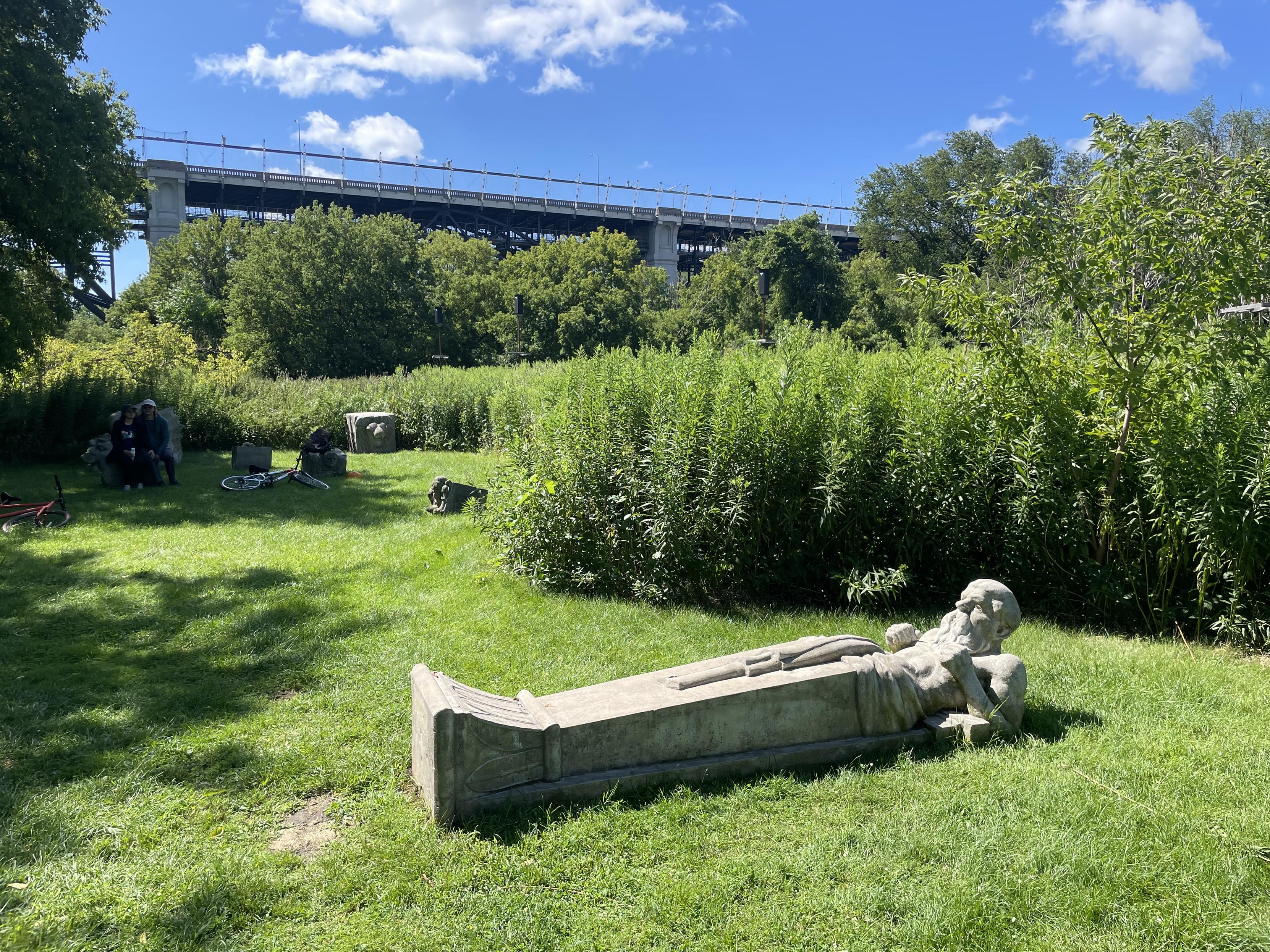 Photo facing south of a green, wooded area.  In the foreground is a stone or concrete figure of a stylized man lying on his back and appearing to  do an ab crunch.  In the background is a bridge carrying cars and a subway on the lower deck.