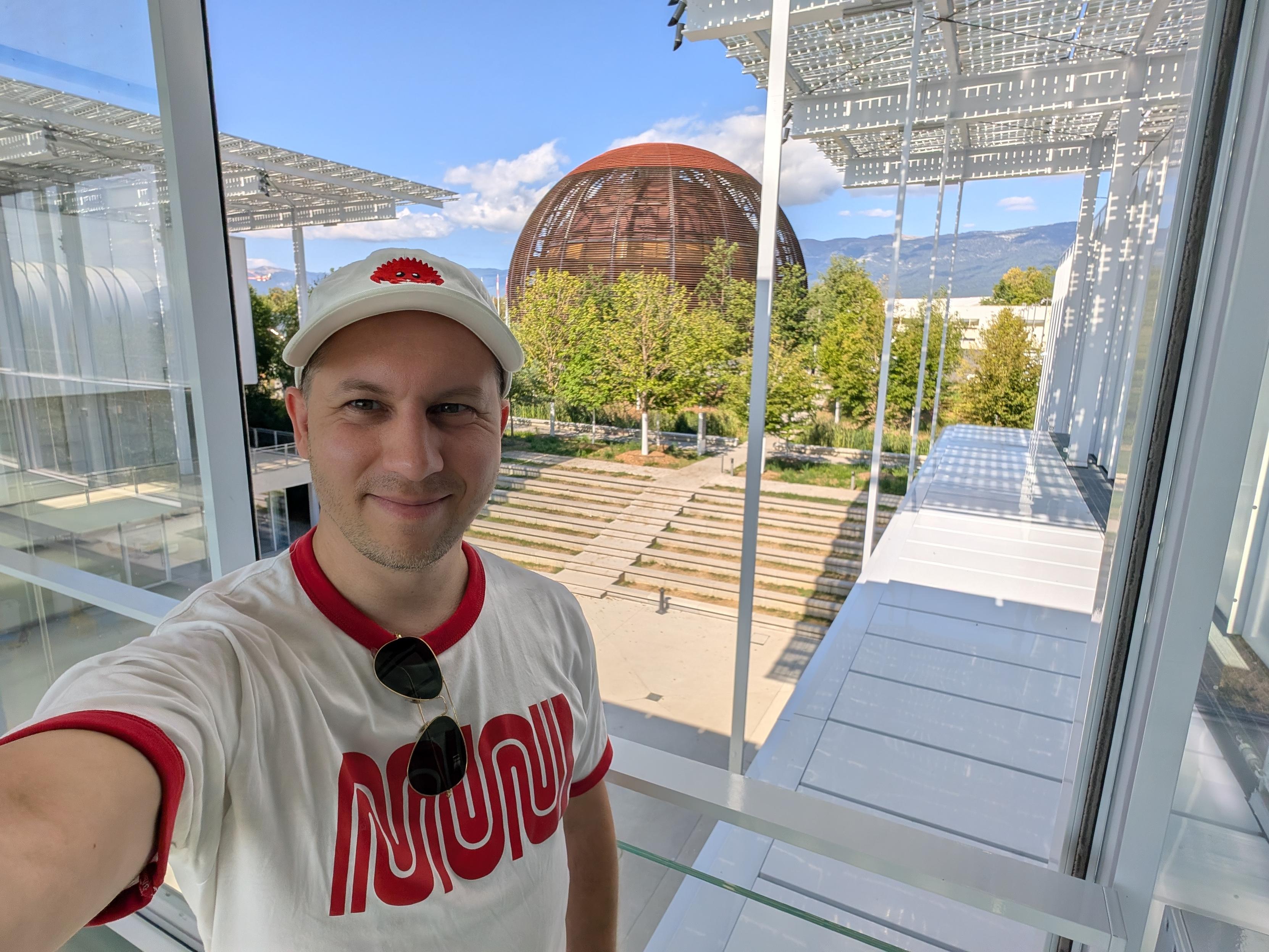 Selfie from a pedestrian bridge overlooking a rusted slates spherical building "Globe of Science and Innovation". I'm wearing a white shirt with the stylized Muni logo in red, and a white cap with the red Ferris crab.