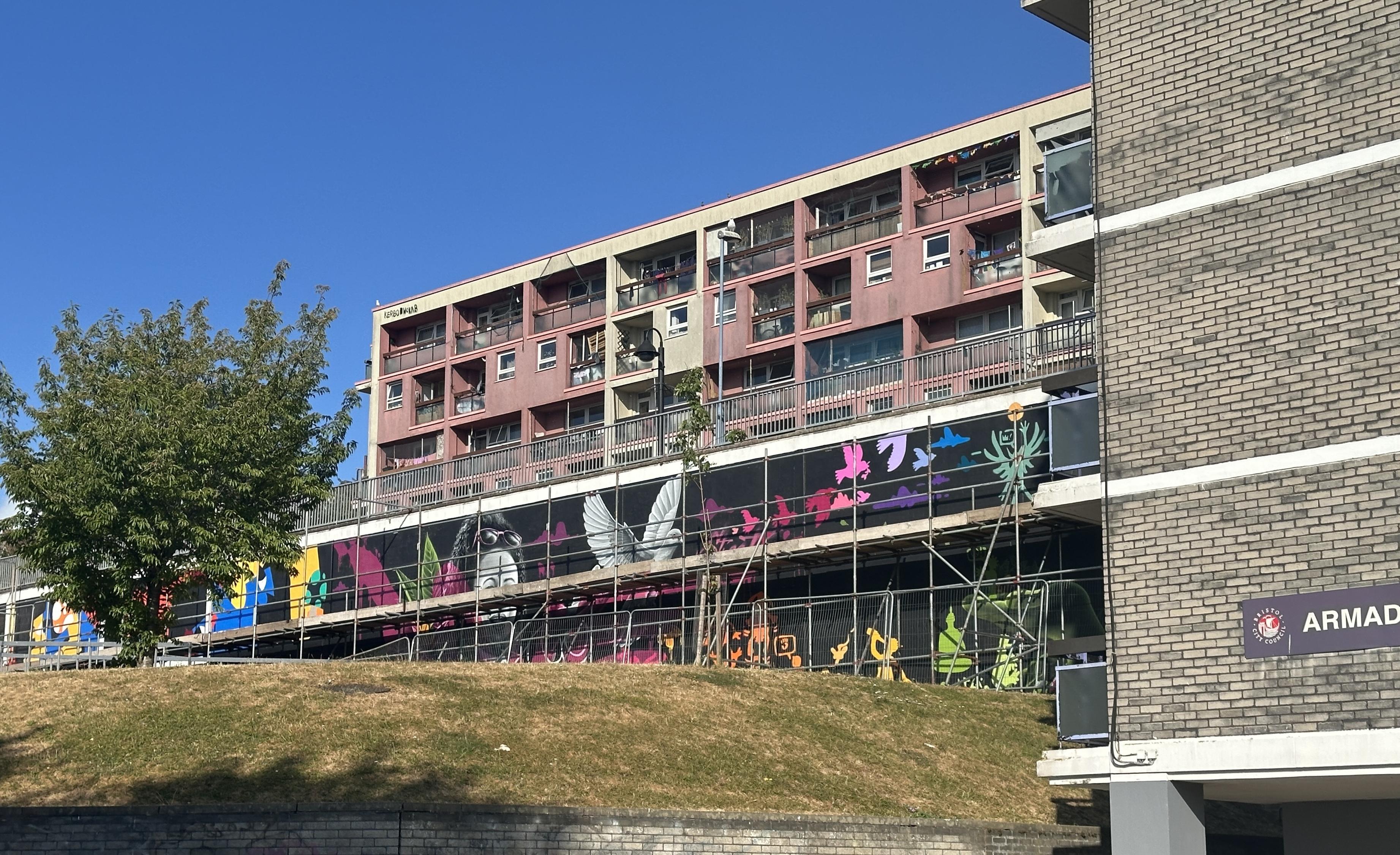 Four storey tower block with murals being painted in front; scaffolding obstruct the murals. They are on a black background and art of various colours. A dove is a central theme