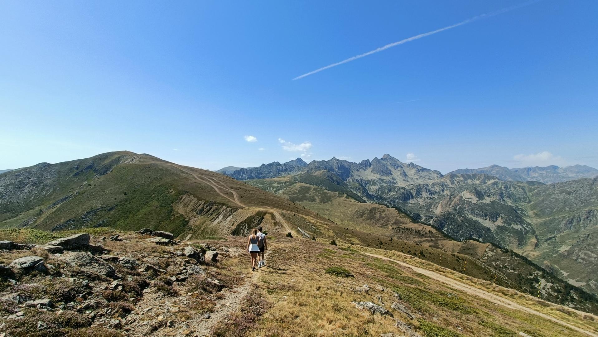 On a ridge path at an altitude of 2000m, 2 people seen from behind are walking. The sky is blue, the slopes alternate grey and green
