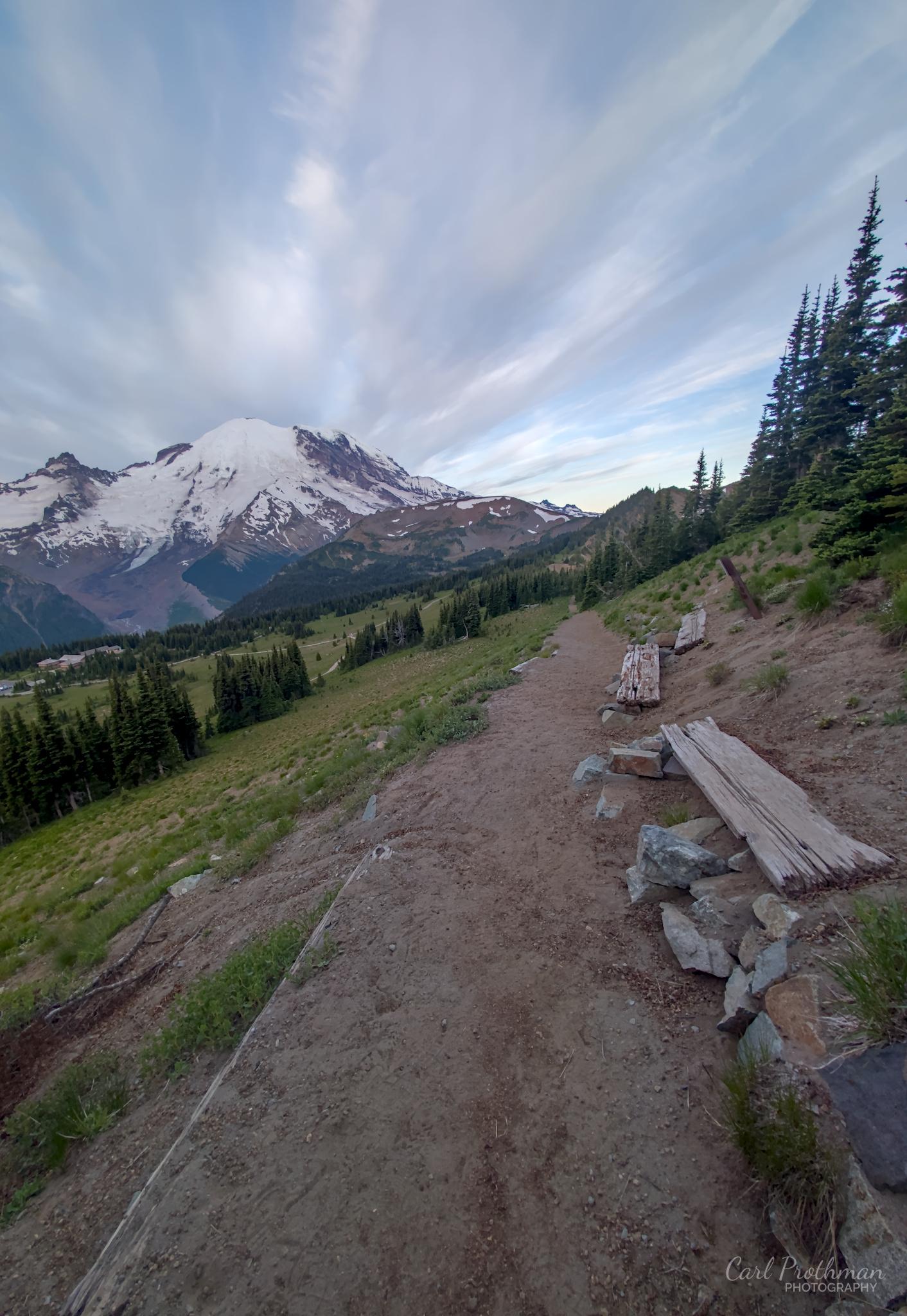 A dirt hiking trail curves along an alpine hillside toward snow-covered Mount Rainier. Weathered wooden benches line the right side beneath evergreens, while long, wispy clouds sweep dramatically across a pale blue sky. The scene feels open, quiet, and inviting.