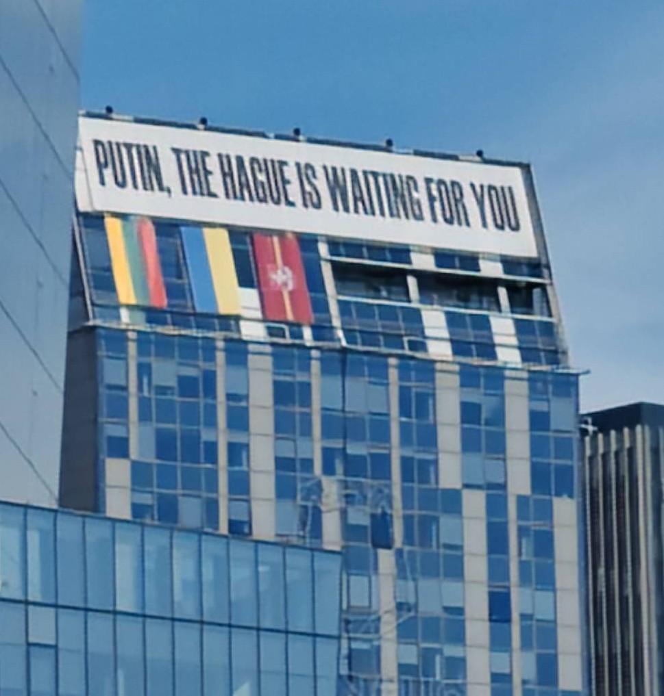 Large text reading, "Putin, The Hague is waiting for you." Below are three flags, one of which is the flag of Lithuania and another of which is the flag of Ukraine. Below that is a hard to see outline on the building which is the city symbol of Vilnius.