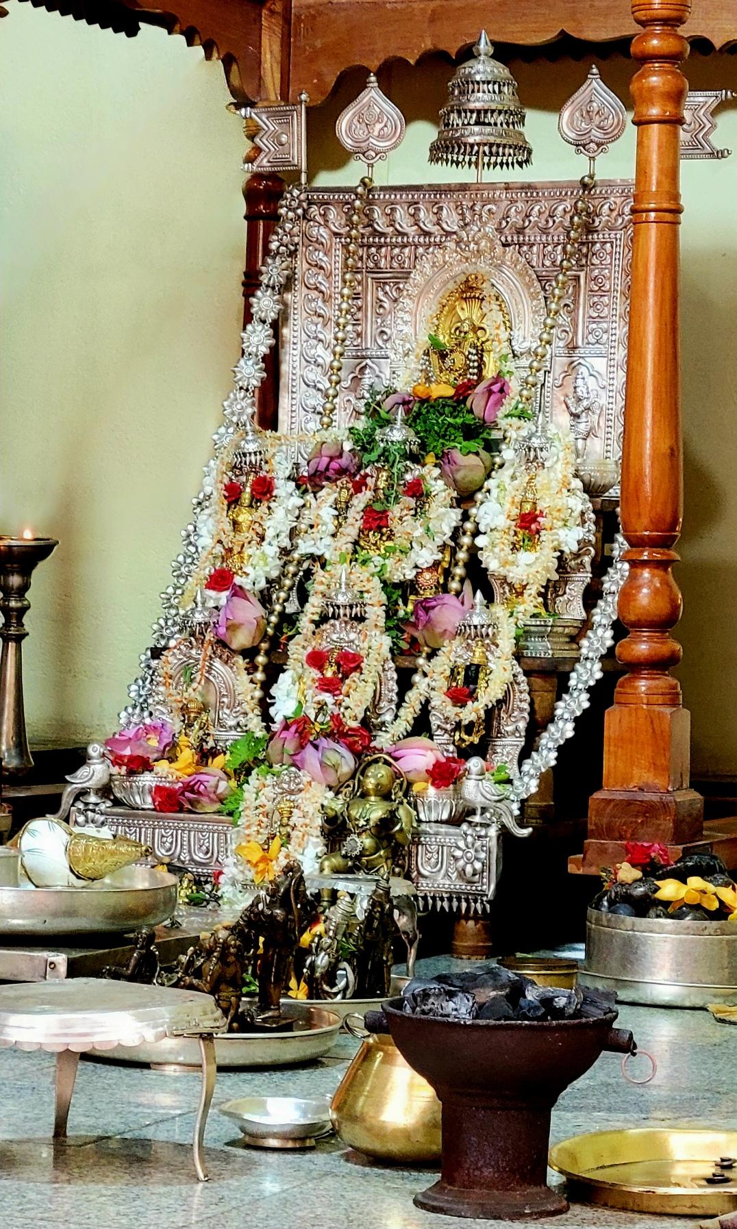 Ornate silver Hindu altar with many bejeweled copper idols which are anointed with loose pink lotuses, jasmine strings, and strings of green holy basil leaves. Other silver and copper idols and utensils used in ritual service are placed in front of the altar.