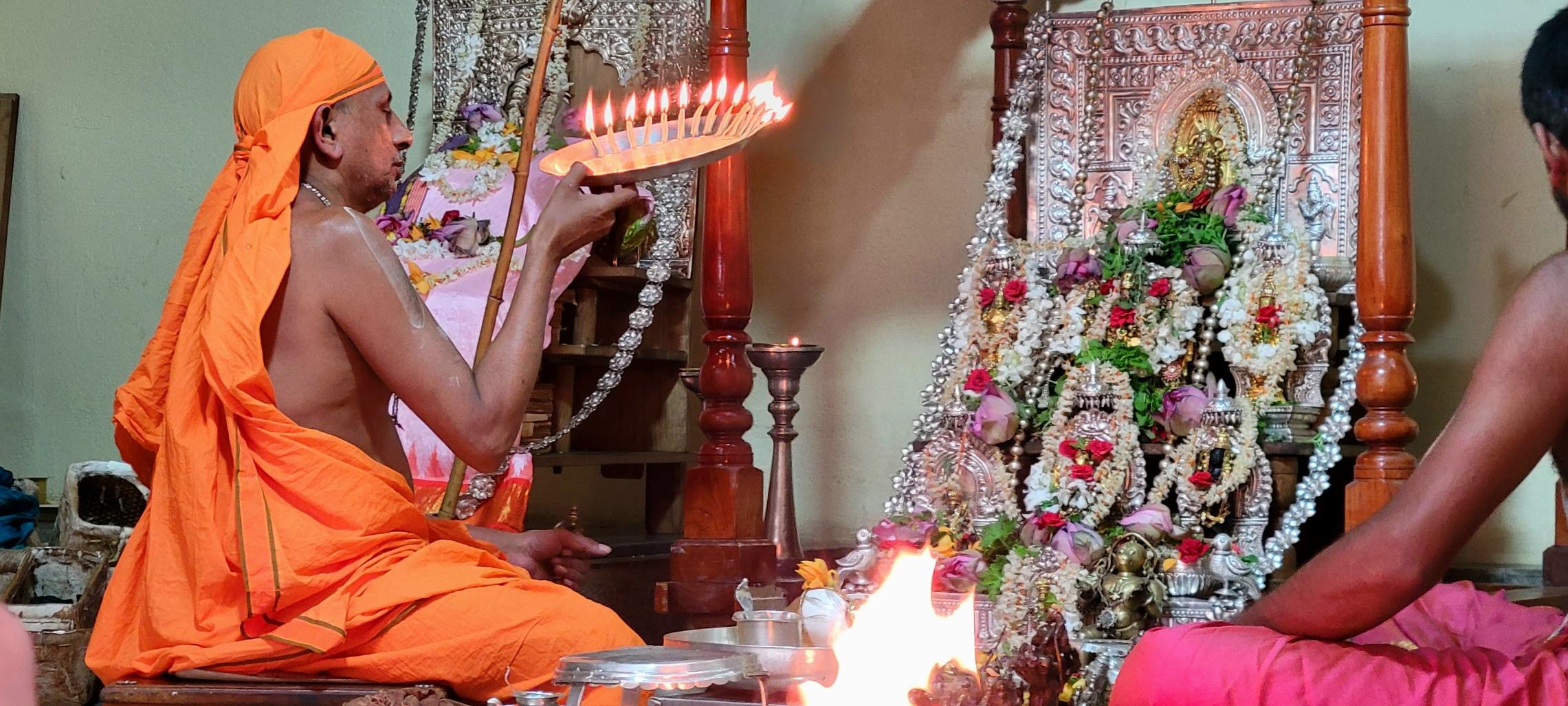 A Hindu Swamiji in saffron robe holds up a silver plate with lit cotton wicks in front of an altar with several idols which are anointed with different  kinds of flowers.