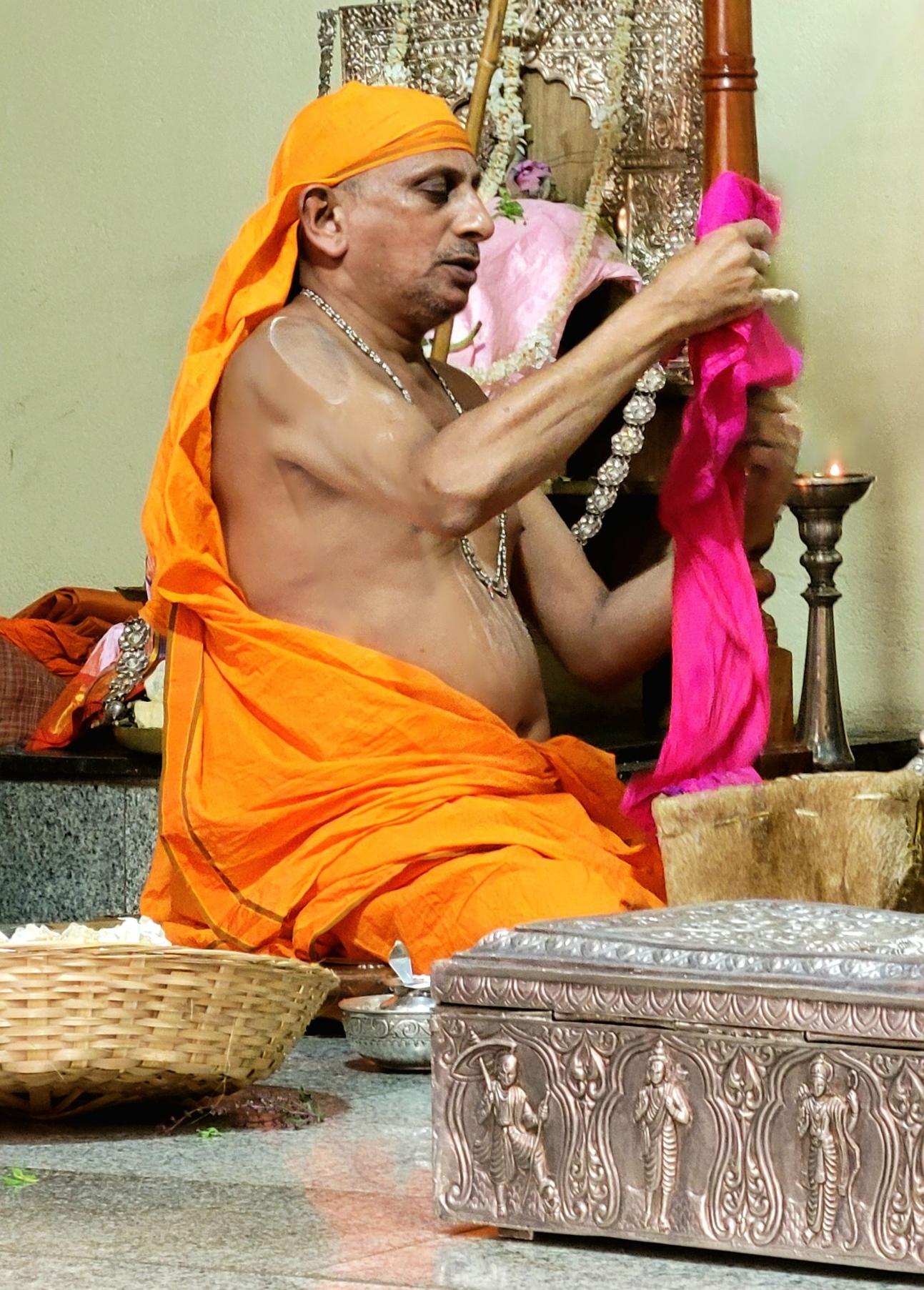 A seated Hindu Swamiji in saffron robe wraps an idol in pink silk cloth.
