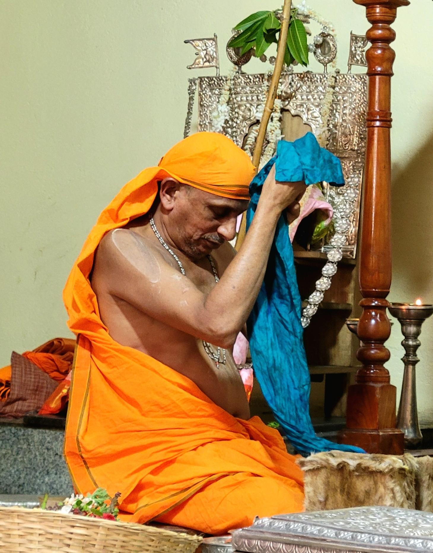 A seated Hindu Swamiji in saffron robe holds to his head an idol that is wrapped in teal silk cloth. Thew tail of the silk cloth hangs down from the Swamiji's hands.