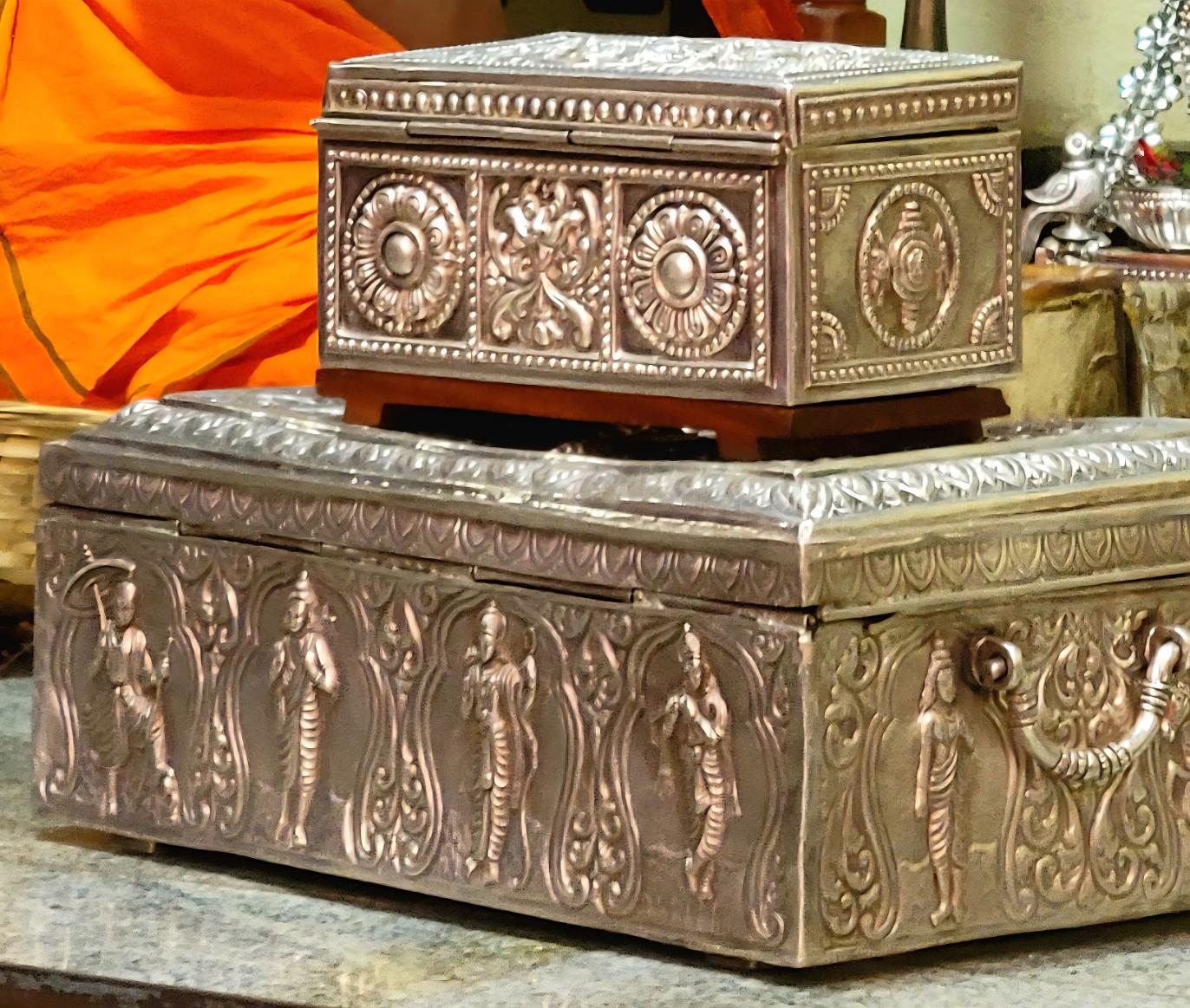 Closeup of a small ornate silver trunk stacked on top of a larger ornate silver trunk. Hindu icons are engraved on the trunks.