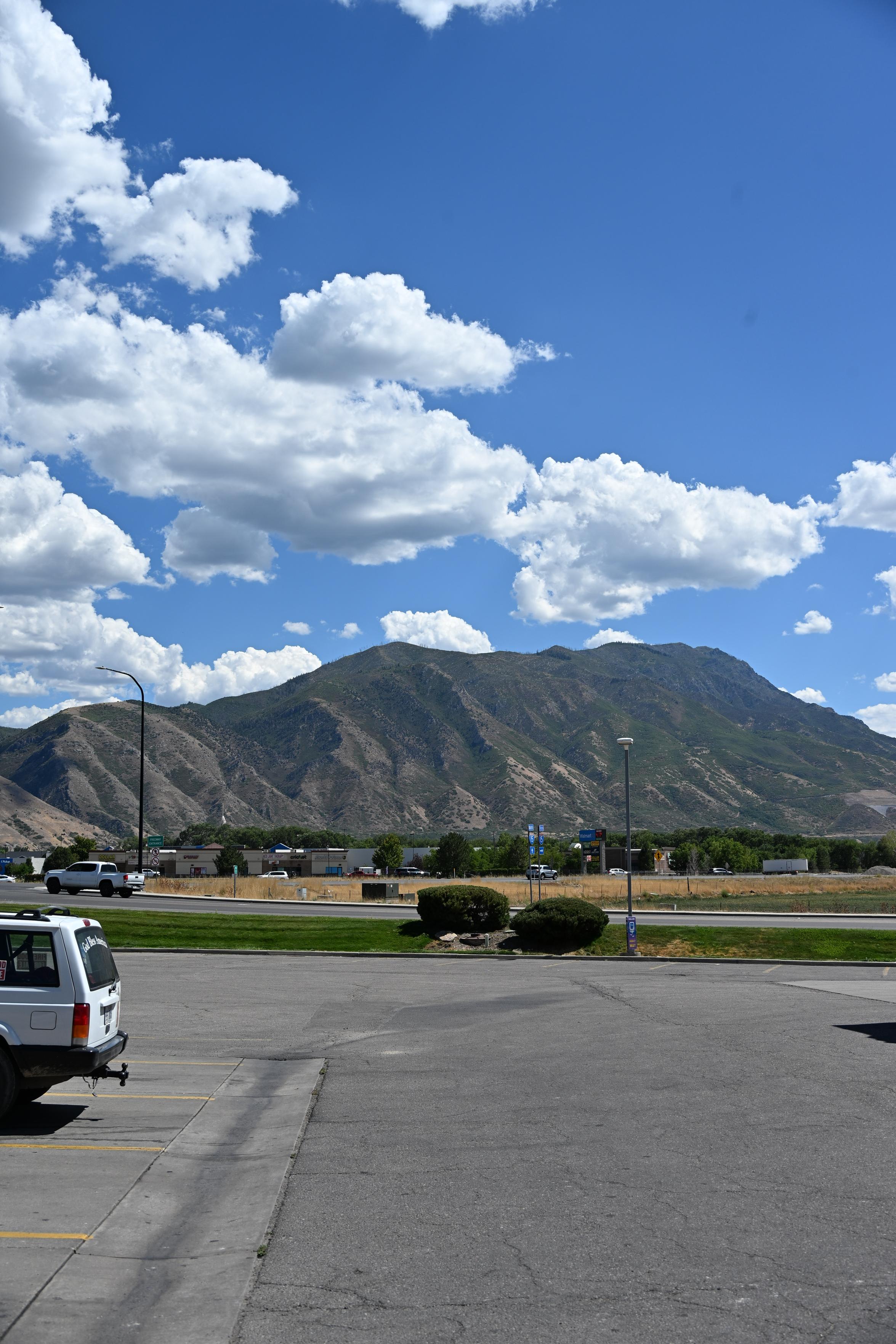 The blue sky is half covered with white puffy clouds spread out. The hills have some green on it, but are otherwise brown. Some green trees are just across the highway. The bottom half is shows the asphalt of a gas station, in rather good condition.