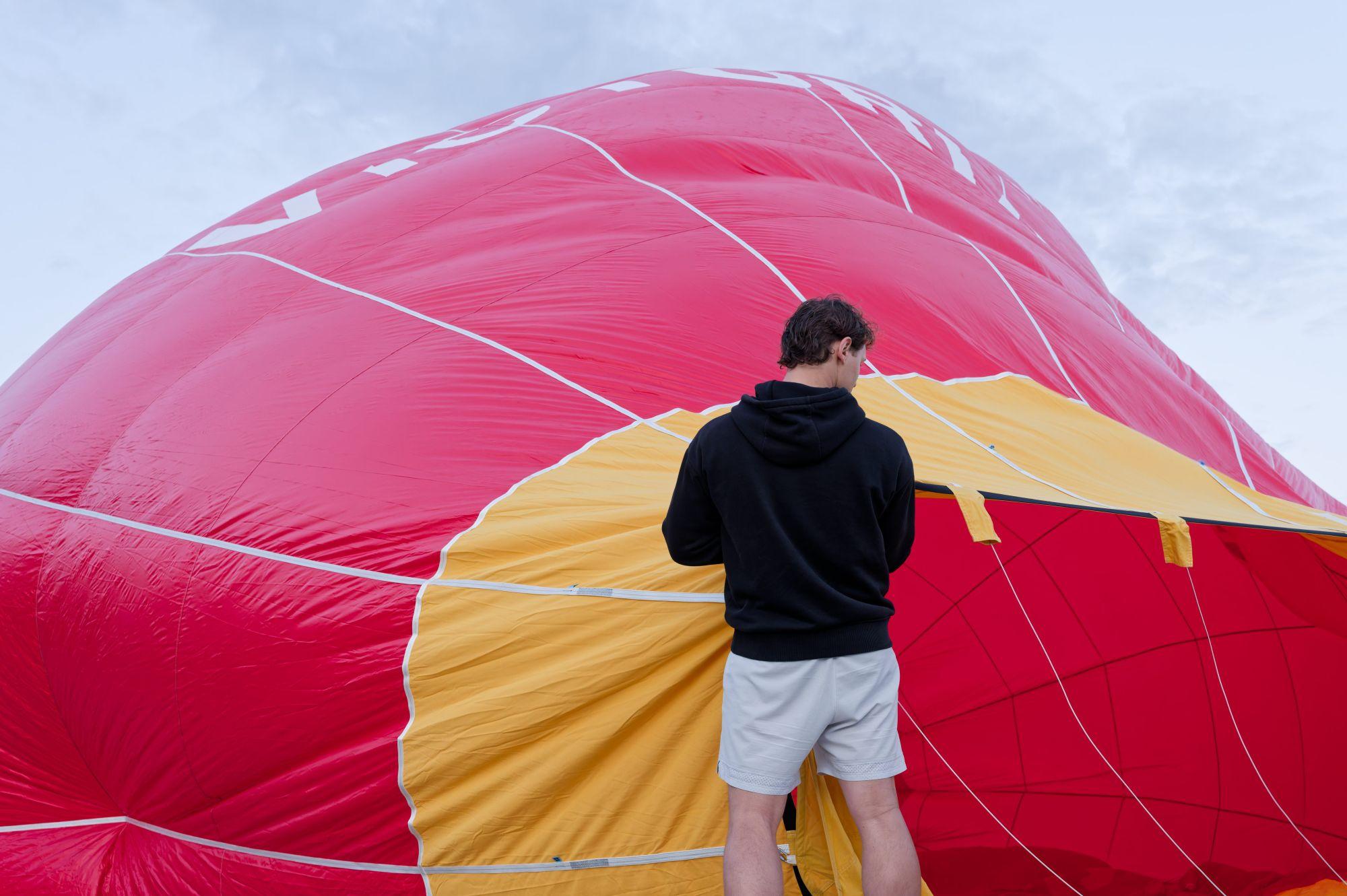 Person in a black hoodie and light shorts stands with their back to the camera, inspecting or adjusting a partially inflated red and yellow hot air balloon envelope spread out on the ground.