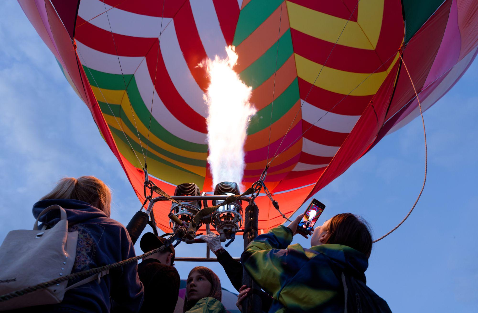 Low-angle view of a crowd gathered around a hot air balloon basket as a bright flame shoots up into the colorful, striped envelope against a twilight sky.