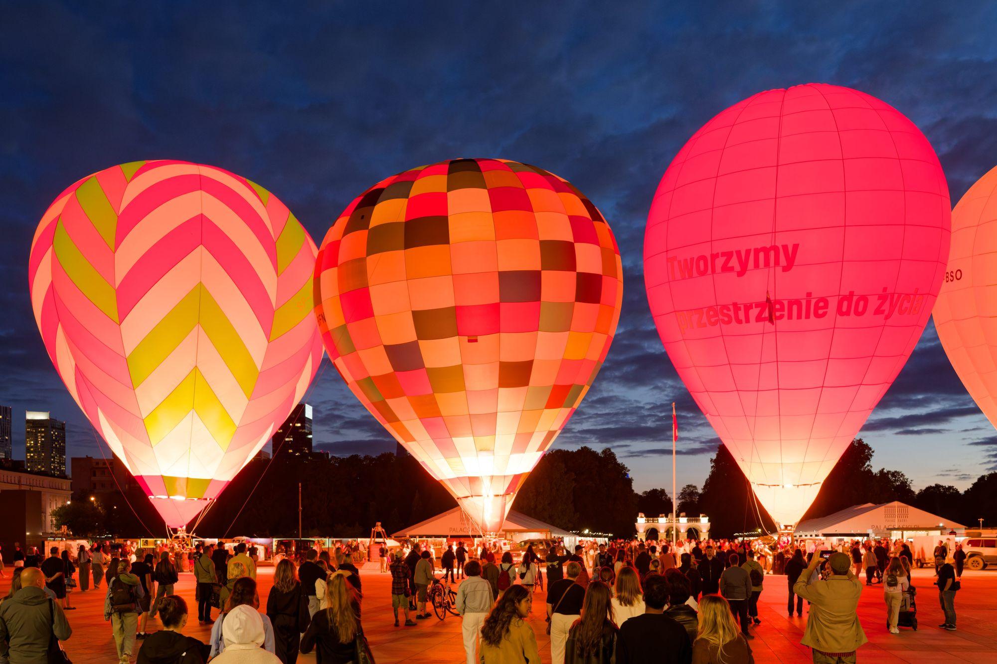 Crowd gathering on Piłsudski Square in Warsaw at dusk to watch several brightly illuminated hot air balloons glowing against the dark sky.