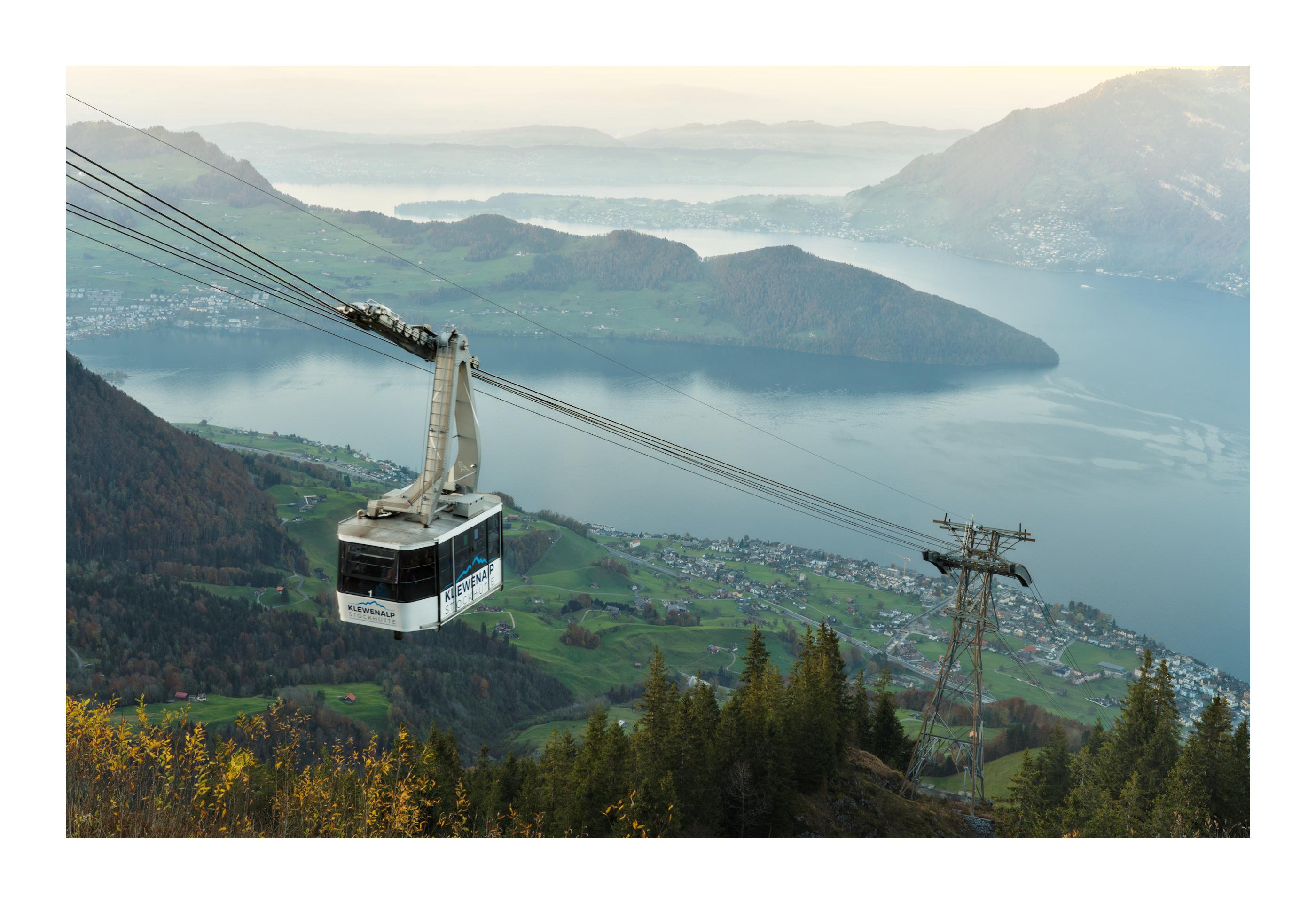 View from a vantage point high above the lake of Lucerne. A cablecar and a pylon are present in the foreground. The village of Beckenried is visible near the lake. The background is composed of rolling hills that bath in the sunset light.