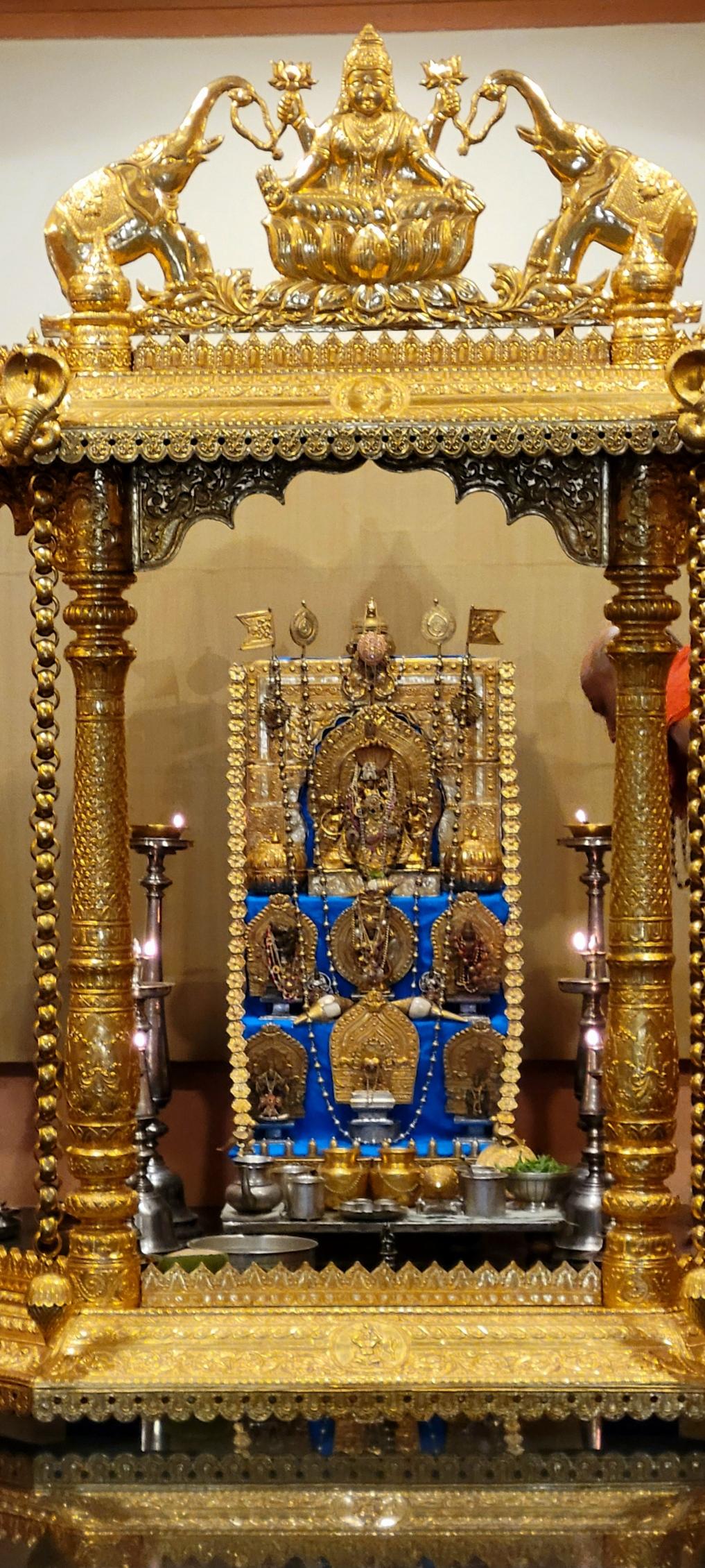 A Hindu altar behind a gilded pavilion. The altar is stepped with multiple idols at each step. A garland of gold flowers on the altar. Icons, utensils, and other ritual objects around the altar.