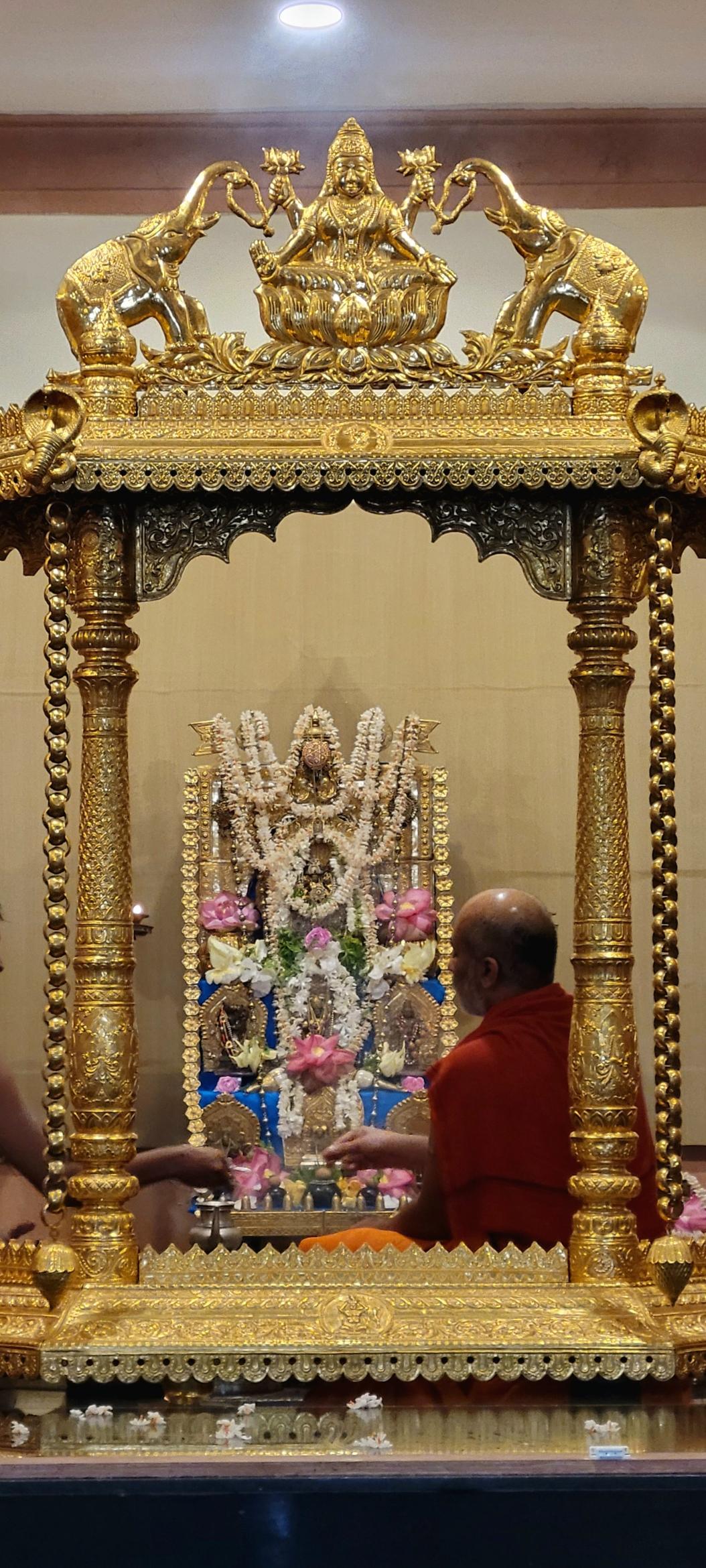 A Hindu altar behind a gilded pavilion. The altar is stepped with multiple idols at each step. A garland of gold flowers on the altar. Strings of jasmine and individual pink lotus flowers on the idols. Icons, utensils, and other ritual objects around the altar. A Swamiji (bald man in saffron robe) sits in front of the altar with his back to the camera.