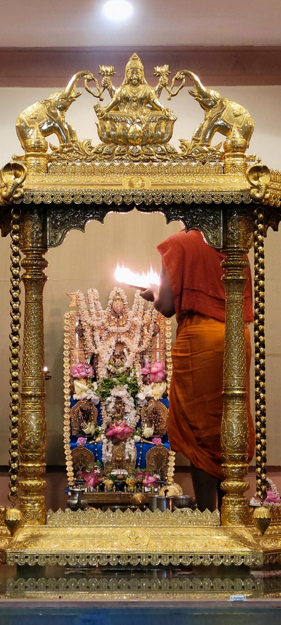 A Hindu altar behind a gilded pavilion. The altar is stepped with multiple idols at each step. A garland of gold flowers on the altar. Strings of jasmine and individual pink lotus flowers on the idols. Icons, utensils, and other ritual objects around the altar. A Swamiji (man in saffron robe) stands in front of the altar with his back to the camera, holding up a plate with several brightly lit wicks and shows the flame to the idols.