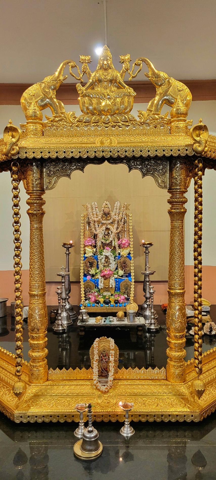 A Hindu altar behind a gilded pavilion. The altar is stepped with multiple idols at each step. A garland of gold flowers on the altar. Jasmine strings and pink lotuses on the altar. Icons, utensils, and other ritual objects around the altar. Three pairs of lit traditional oil lamps in front of the altar.