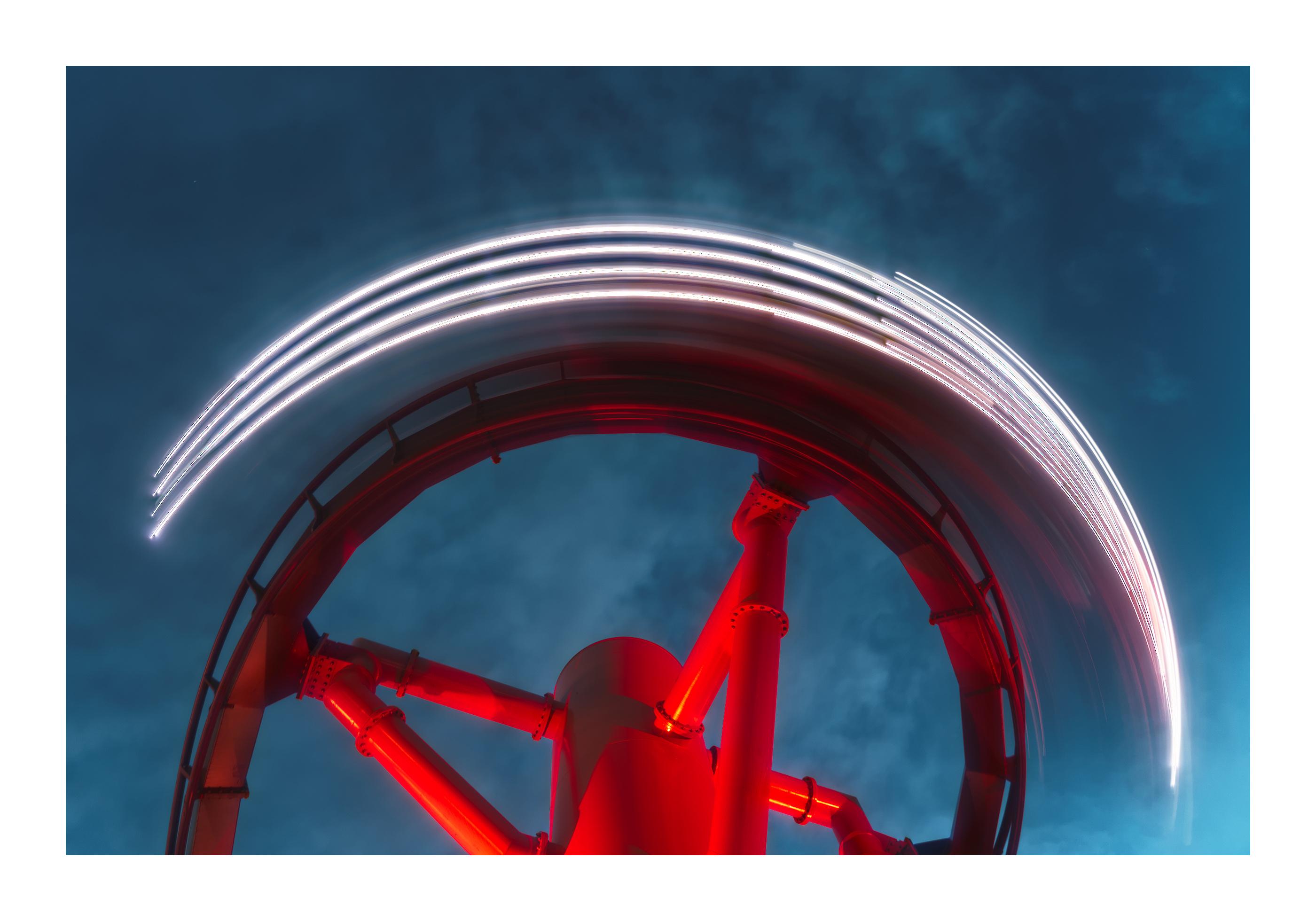 Long exposure (0.5 second) photograph of the inverted roller coaster "Monster" at Gröna Lund, taken during blue hour. The orientation is intentionally confusing (no horizon is present on the picture). The lights on the roller coaster train are describing a half circle. The roller coaster track is illuminated with a bright red light, contrasting heavily with the blue sky that compose the entirety of the background.