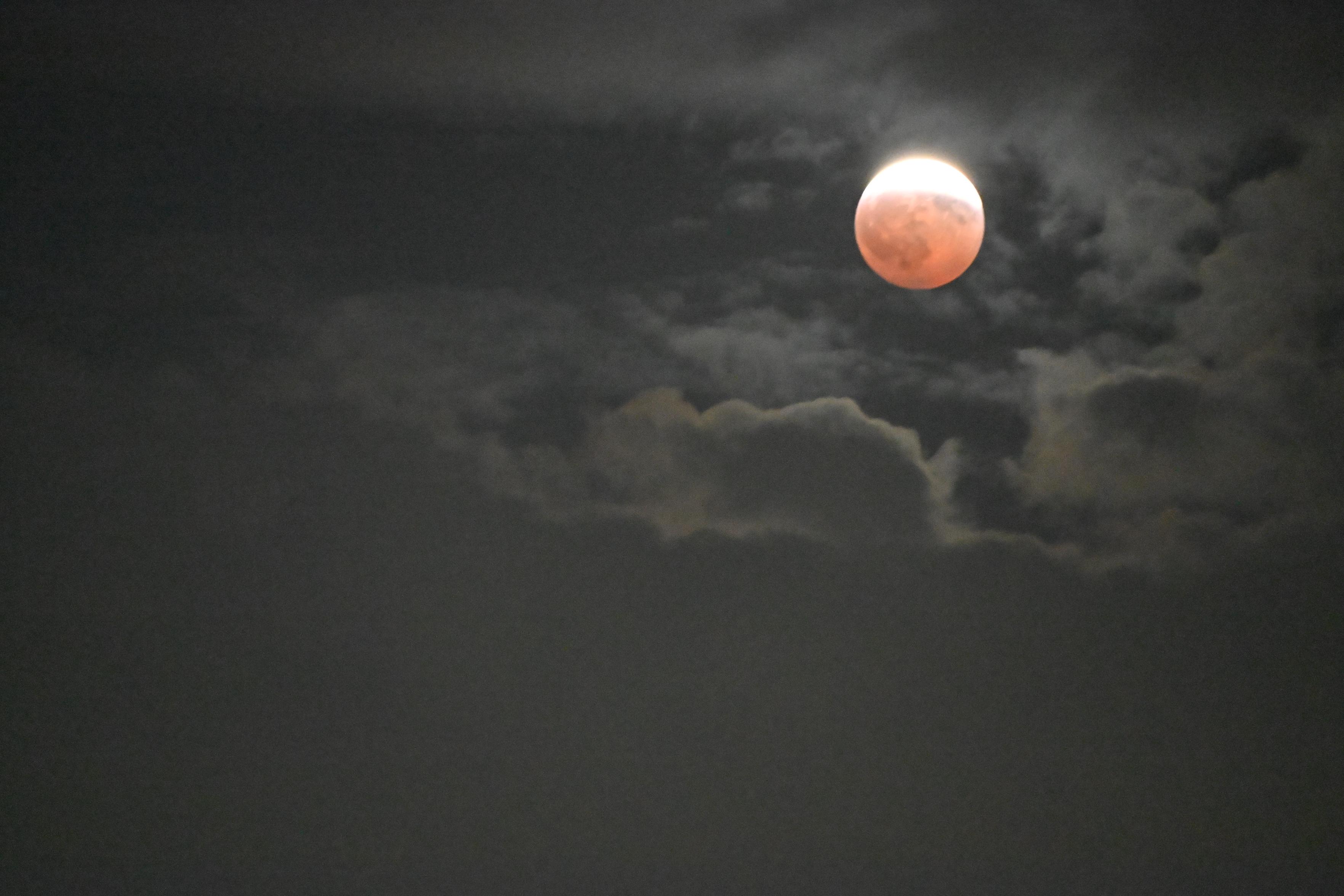 A partial eclipsed moon. We can see the craters on the red/obscured part. The still lit part is washed out in white. Clouds are above and below the moon, showing the narrow band of time to get the shot.