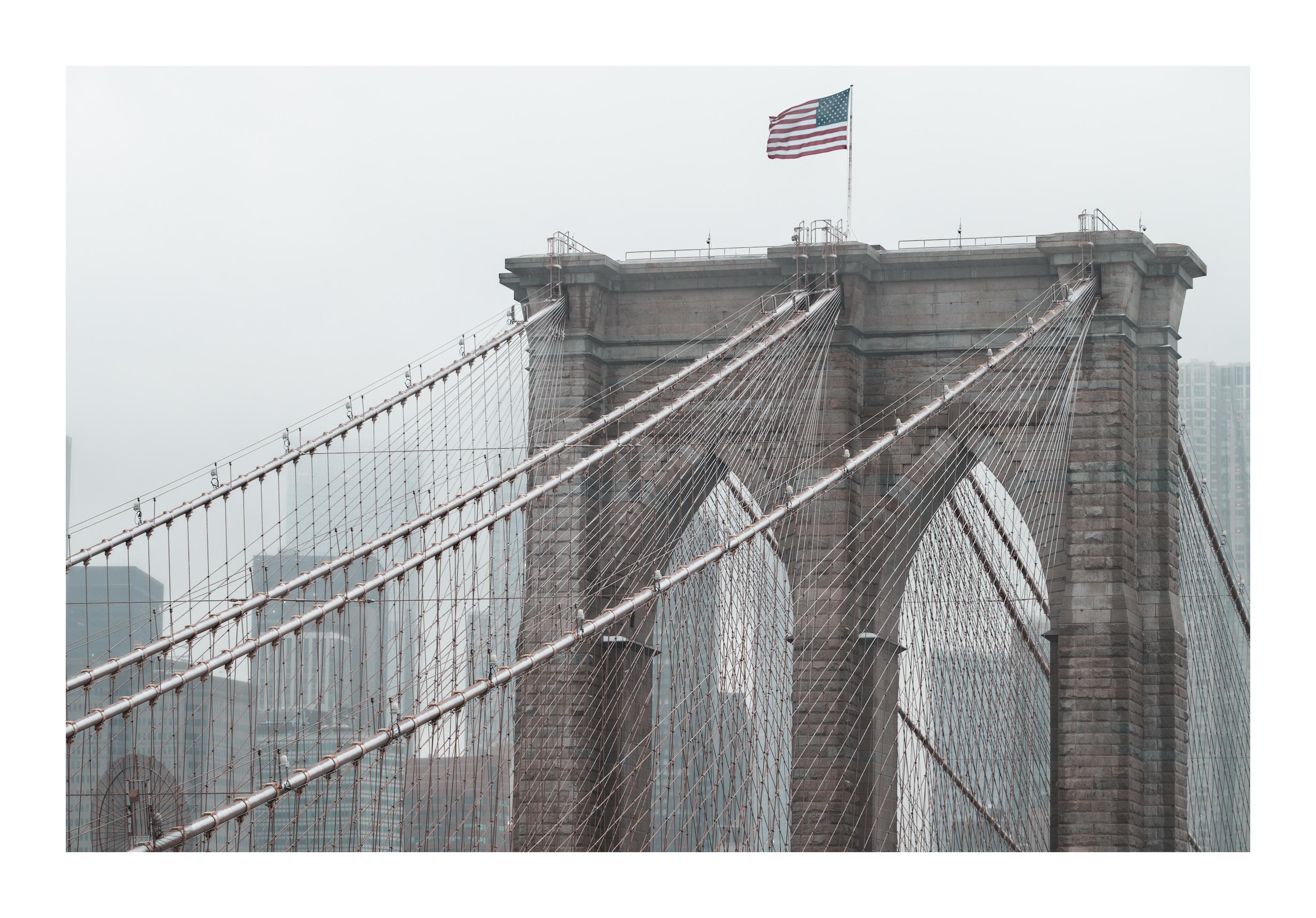 The Brooklyn Bridge is partially seen with a touch of snow on its main suspending cable. The 2 Gothic-style arch windows of one tower are visible but the other tower, as well as the main span isn't. The background is composed of the downtown Manhattan skyline, melting in the high fog. Buildings are not easily recognisable.