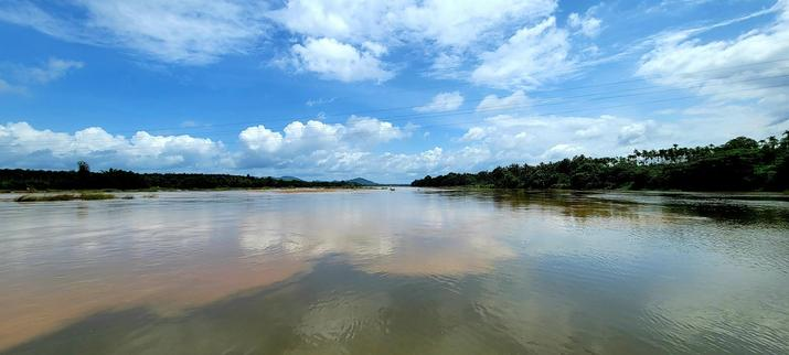 Wide angle shot of two rivers meeting. Screen left, the water is red due to dirt from fresh rains. Screen right, the water is green. Green tree-lined banks on both sides of the river(s) (and the combined river is very wide). Bright blue sky with some white clouds. Sunlight highlights the clouds as well as parts of the river water.