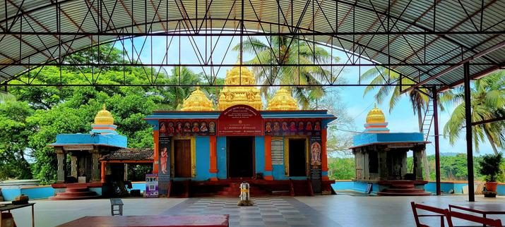 A Hindu temple on a river bank. Two small buildings on the side, larger one in the middle. The middle building has three cupolas; the other two buildings have one cupola each. All buildings are painted blue and red. The cupolas are painted gold. A large tiled yard with a roof overhead in front of the buildings. Green trees in the background.