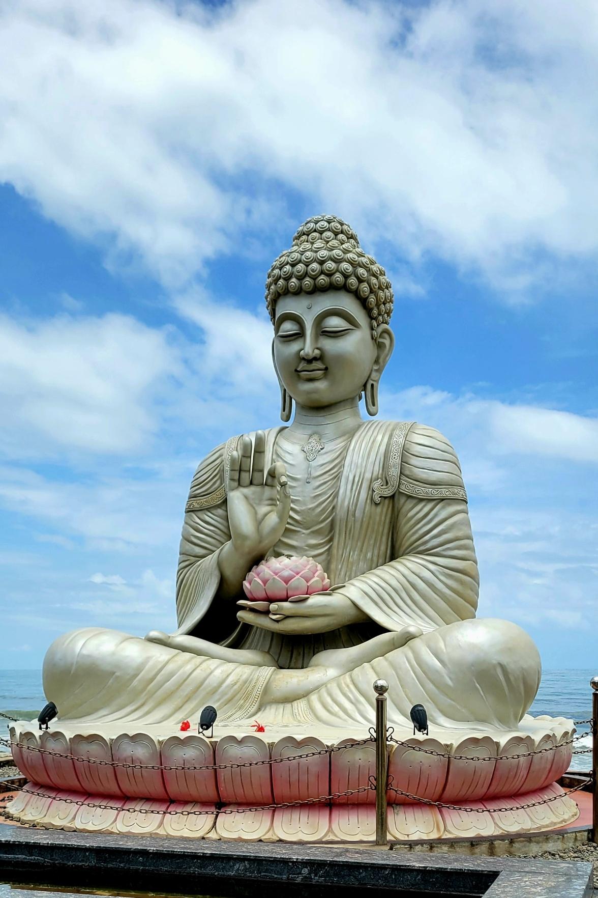 Closeup of a large installation of a silver Buddha seated inside a pink lotus. Holding right hand in meditative posture; a pink lotus in left hand. A sea in the background. Blue skies with some white clouds above.