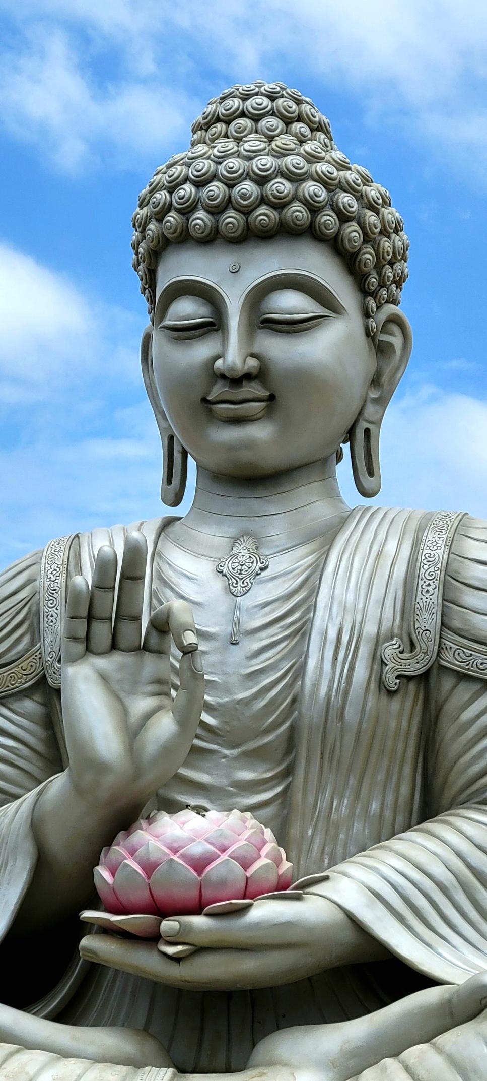 Super closeup of a large installation of a silver Buddha seated. Right hand in meditative posture; a pink lotus in left hand. Blue skies with some white clouds in the background.