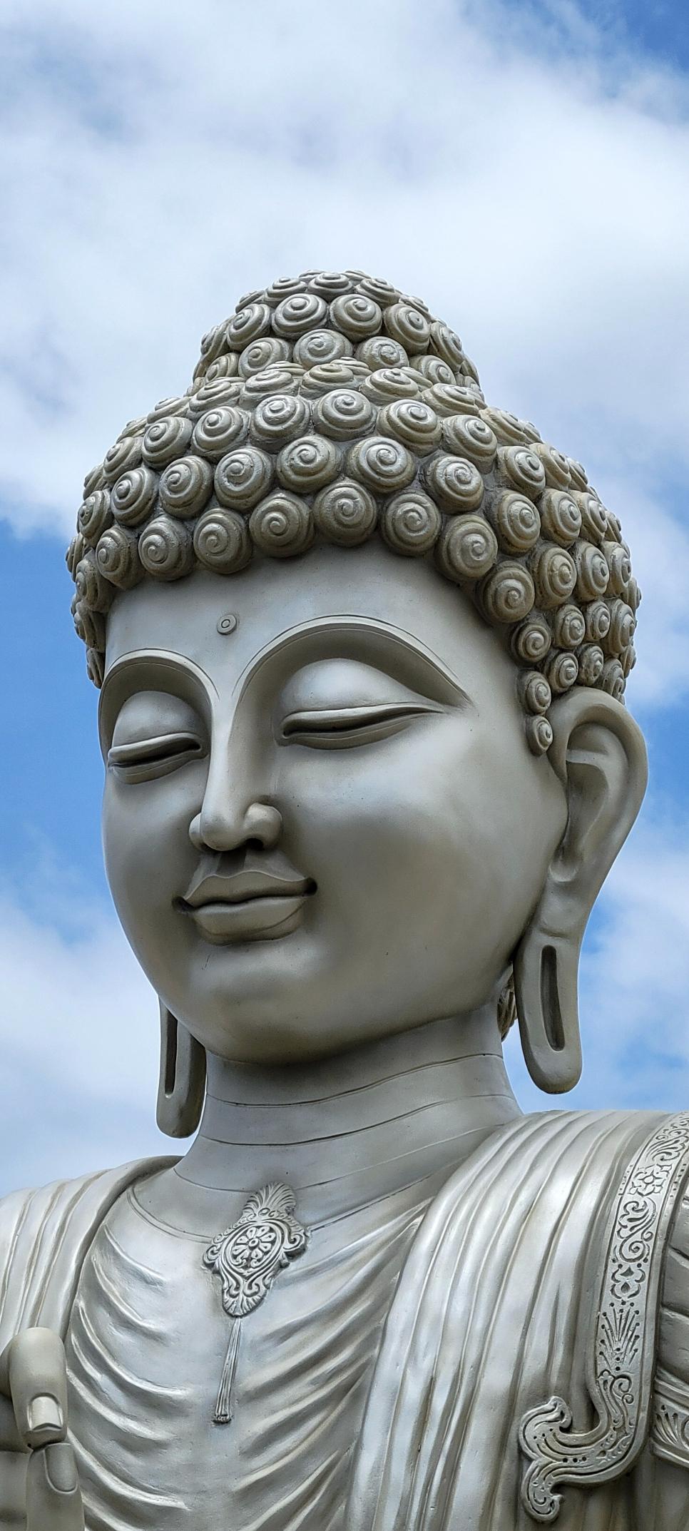 Super closeup of a large installation of a silver Buddha seated. Just the bust is visible. Blue skies with some white clouds in the background.