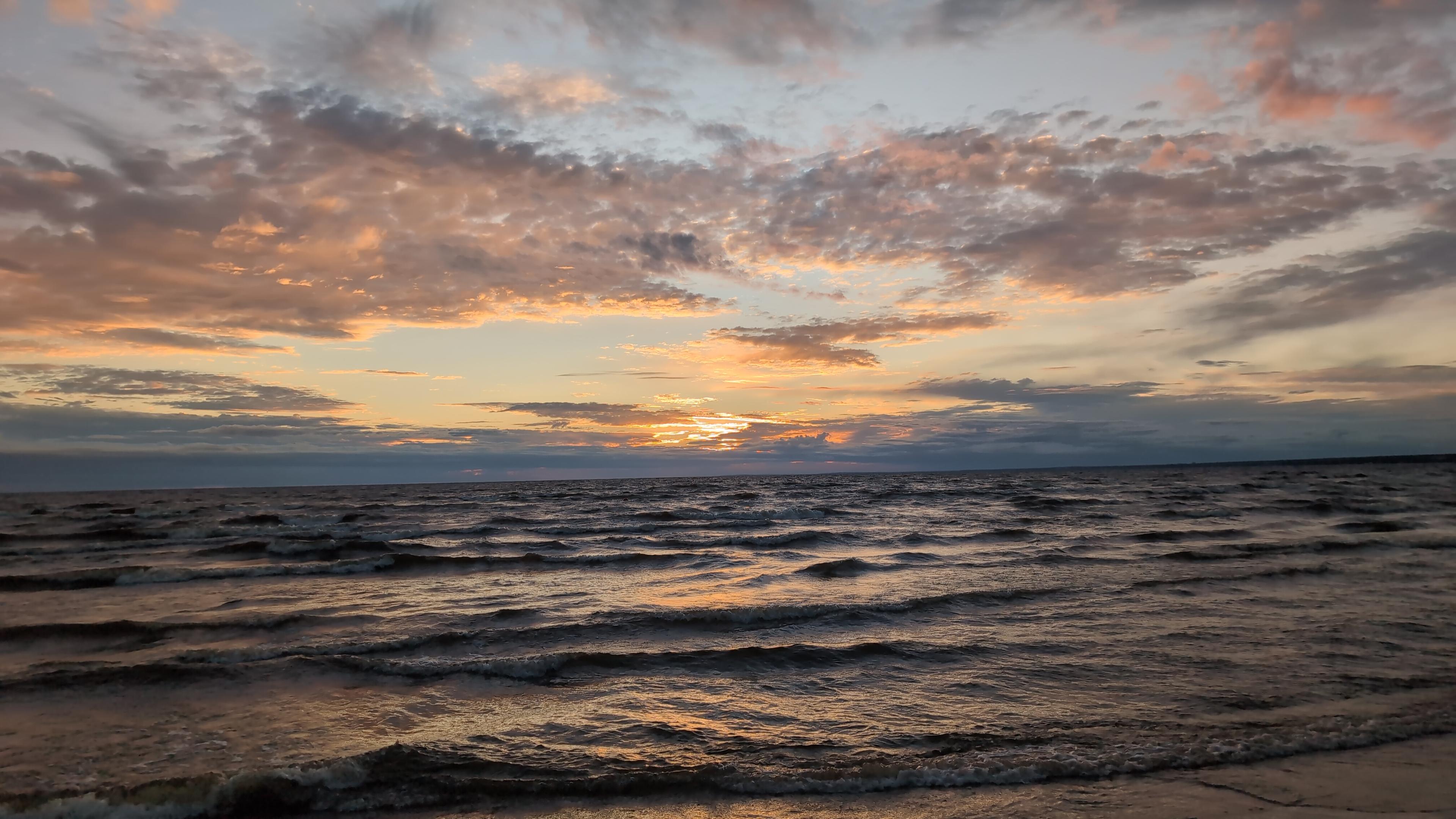 A sunset over the sea full of pastel colours. The sea is heavy and wavy, neither calm nor stormy. Its colour is that dark that it's slightly blue than the wet sand in the right corner, and there's little of orange as a glint. The horizon is a line of grey-blue clouds which the sun into. After a pastel blue gap curly light clouds go with pinky-orange shine. The heavy horizon and the curly clouds shape something resembling a halo.
