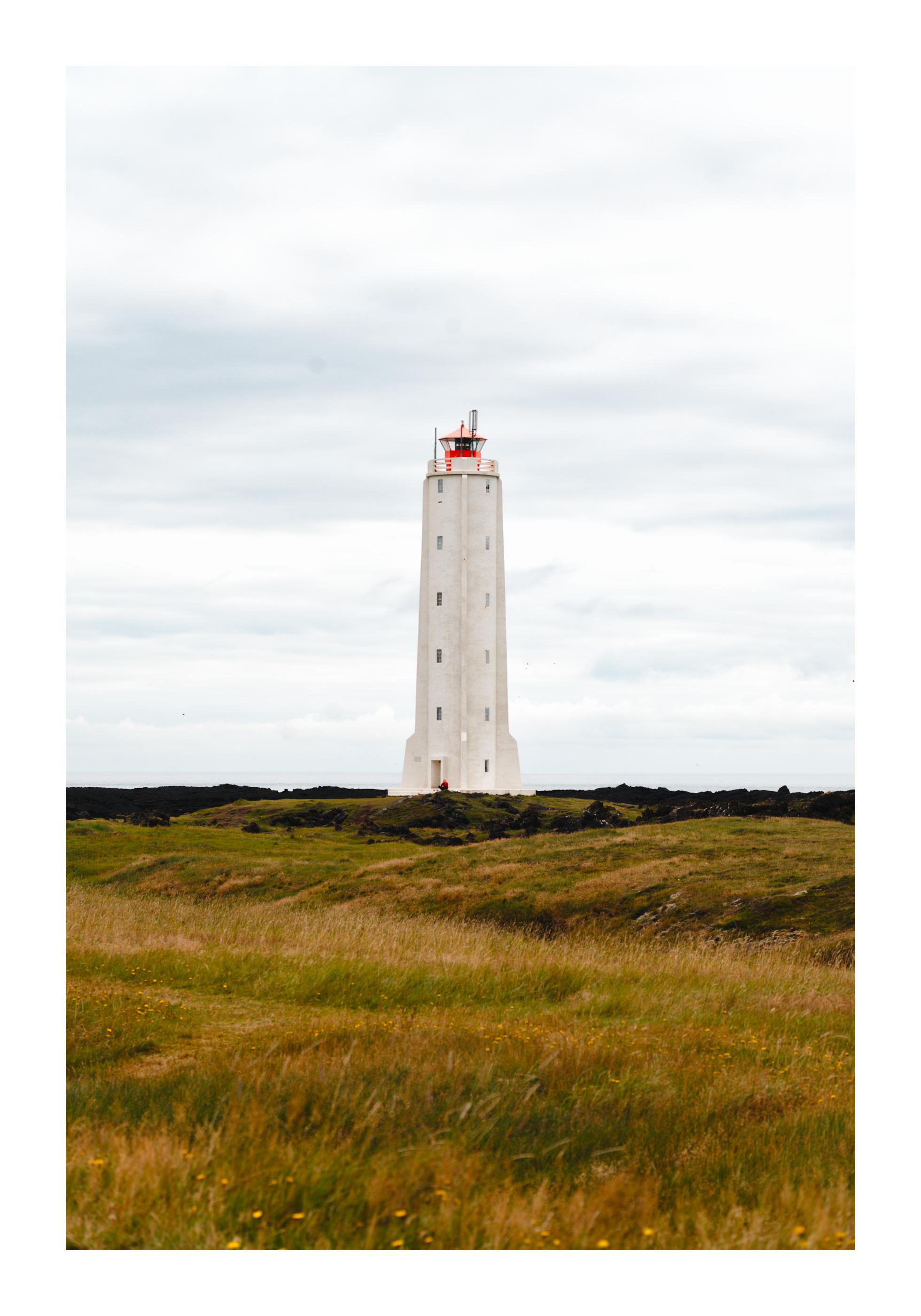 Photograph of a light grey lighthouse topped with a red roof. The ocean is barely visible in the background, and the foreground is composed of small bumps in a green/orange grass. The sky is cloudy and very close in color to the lighthouse itself. The composition is minimalist, with only the sky and grass and lighthouse being prominent. A closer inspection reveals a man with a red jacket sitting at the foot of the lighthouse.