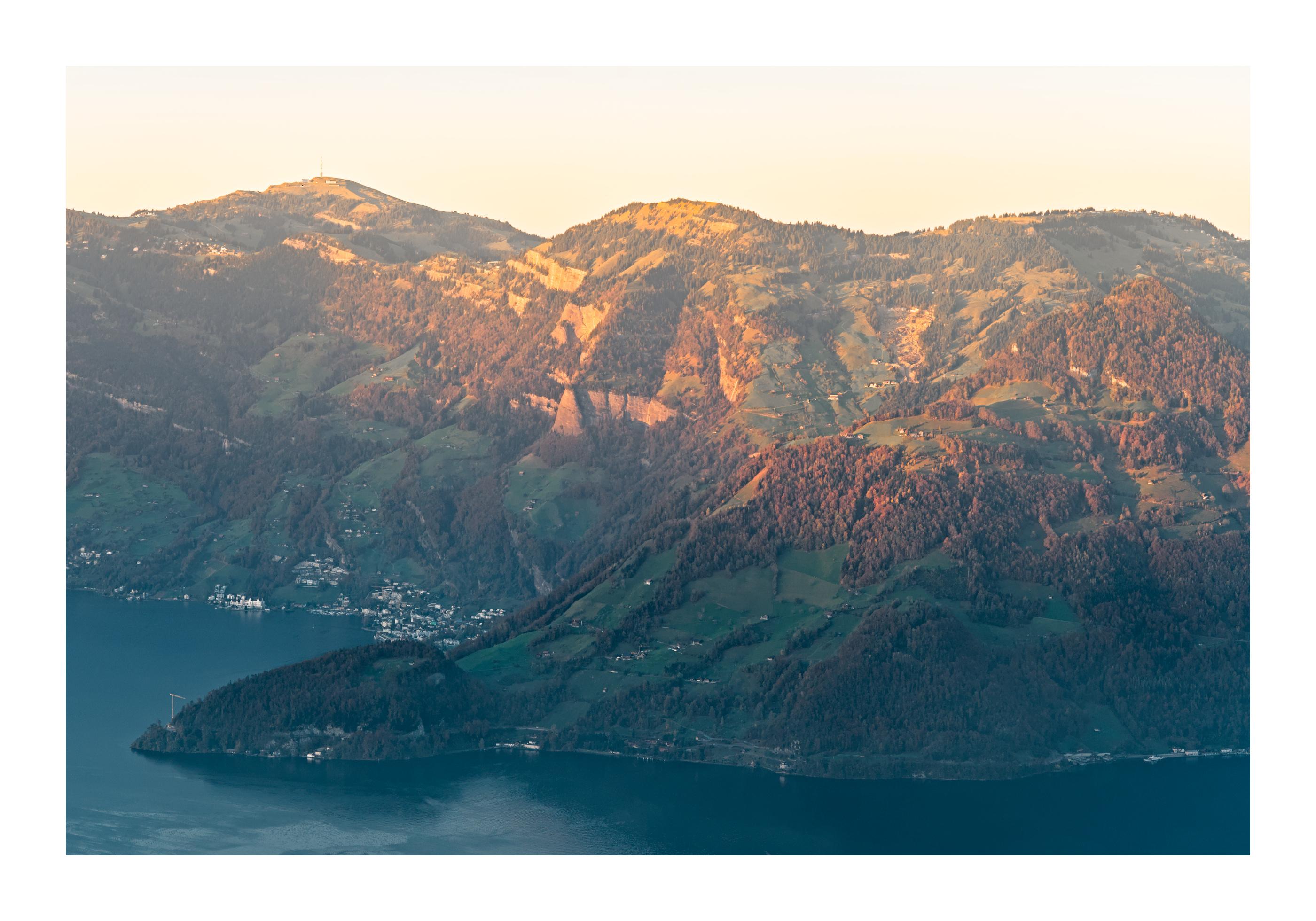 Landscape photograph of a mountain range in Switzerland. A lake is visible at the bottom of the frame, and the top of the mountain is lit by the last rays of sunlight for the day. The colors are bright red/orange there and contrasts with the cooler colors of the bottom of the mountainscape. A village is present at the foot of the mountain by the lake, when zooming in.