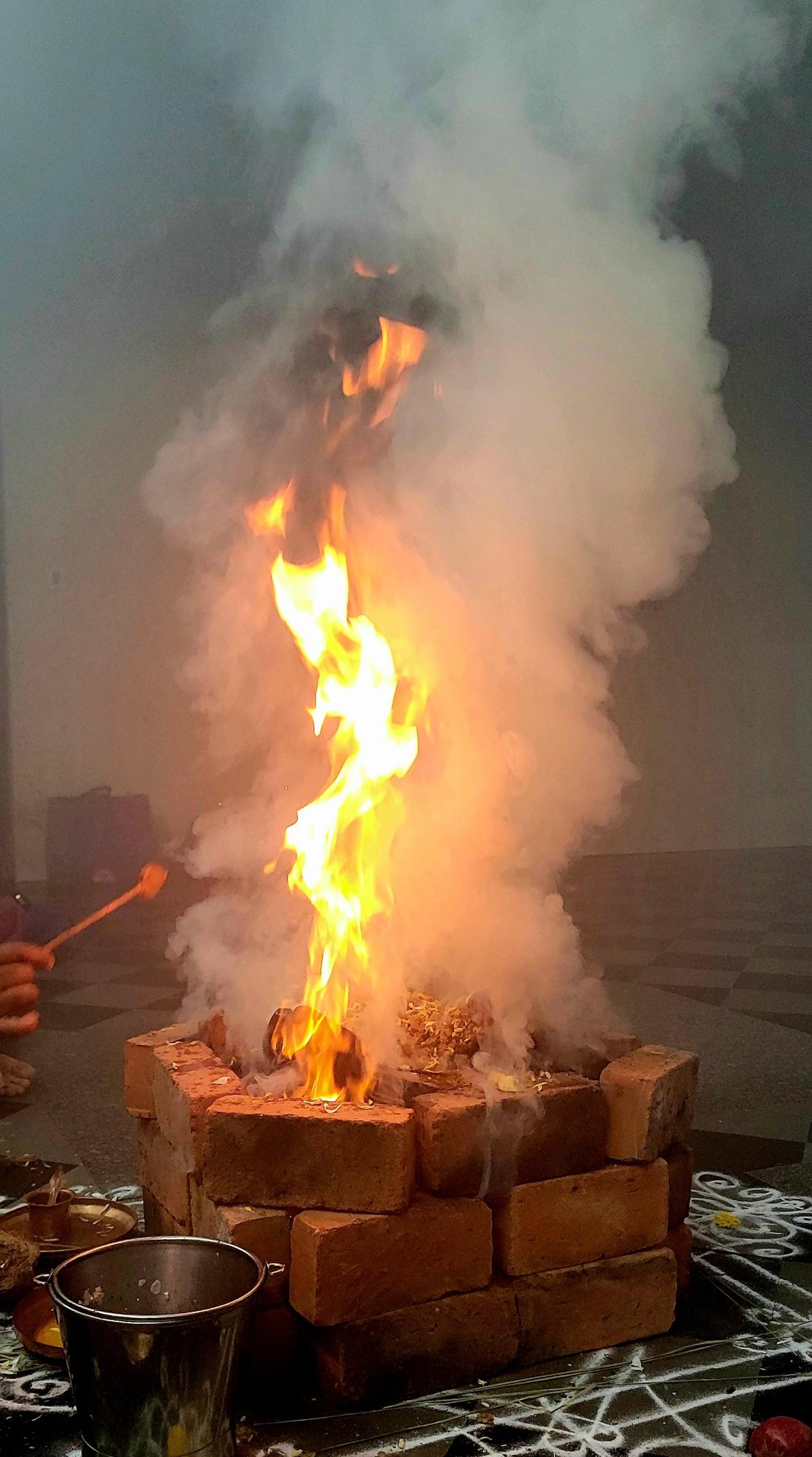 Closeup of a rather high red and white flame coming out a fire pit.  Quite a bit of white and gray smoke around the flame. Objects inside the pit are not discernible. A copper spoon used to add ghee to the pit is visible in someone's hand on screen left.