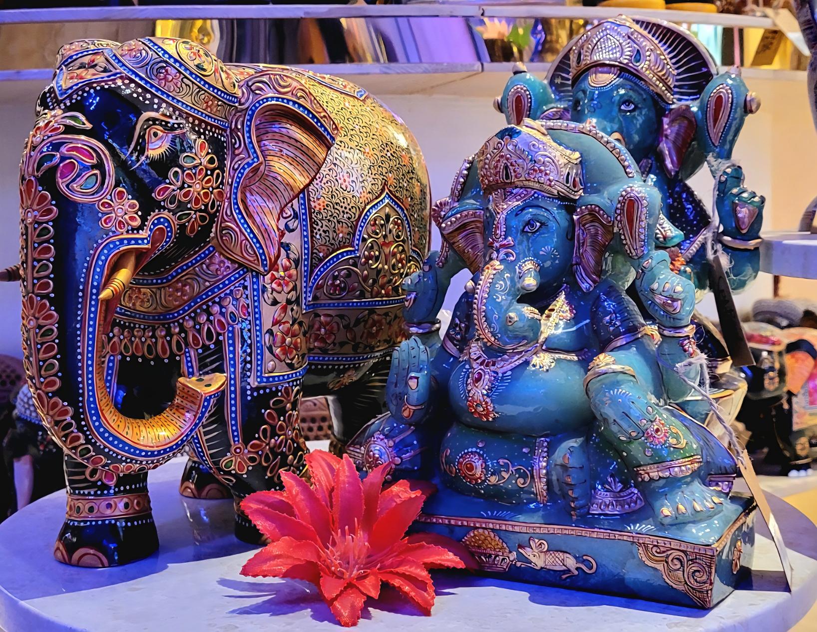 Small sculptures of an ornate black elephant and a blue Ganesha (Hindu deity) side by side at a storefront. Both idols have elaborate painting and embeddings on them. A red flower is placed in front of Ganesha.
