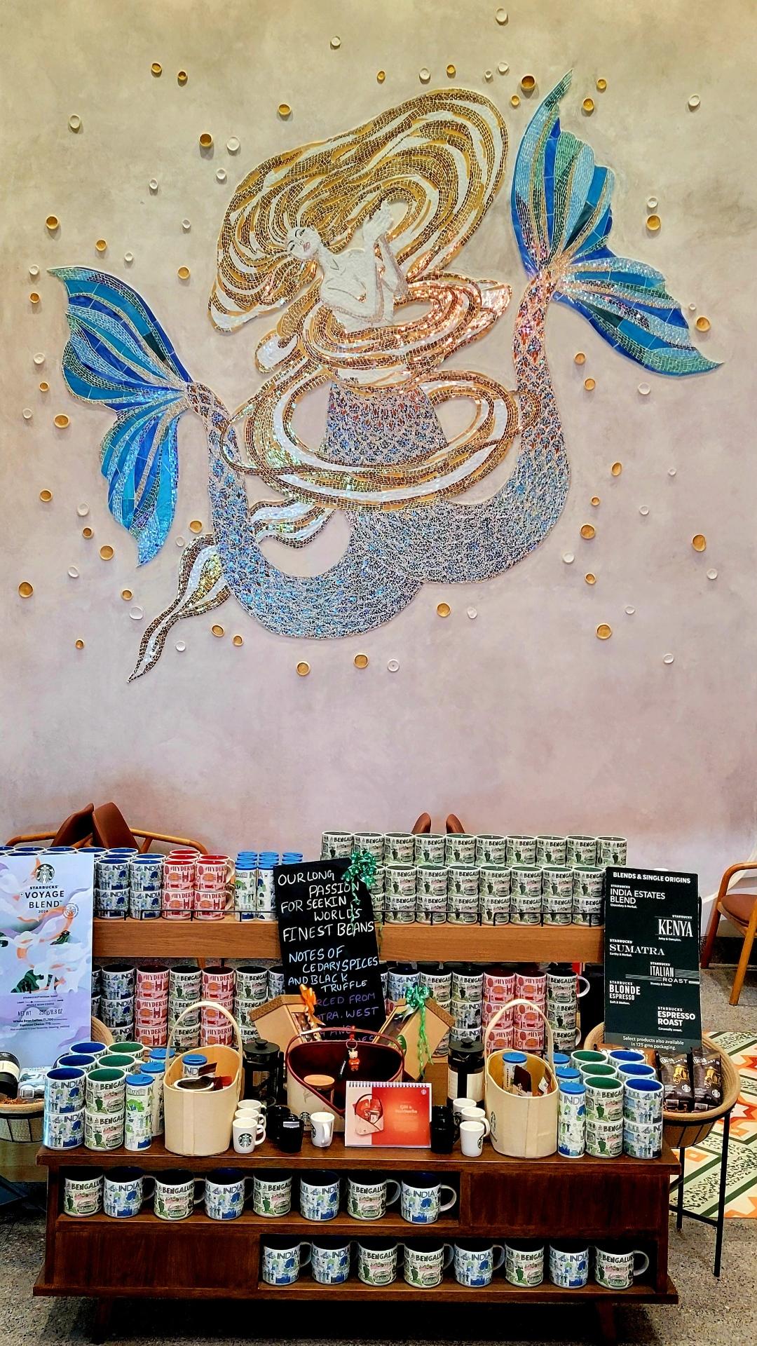 An elaborate rendering of Starbucks's (US coffee chain) siren on a beige-orange wall. The siren's lower body and fins are shades of blue; the upper body is mostly made from lines of gold hues. On the floor, in front of the wall, is a shelf with Starbucks merchandise (mostly mugs) and bags of coffee.